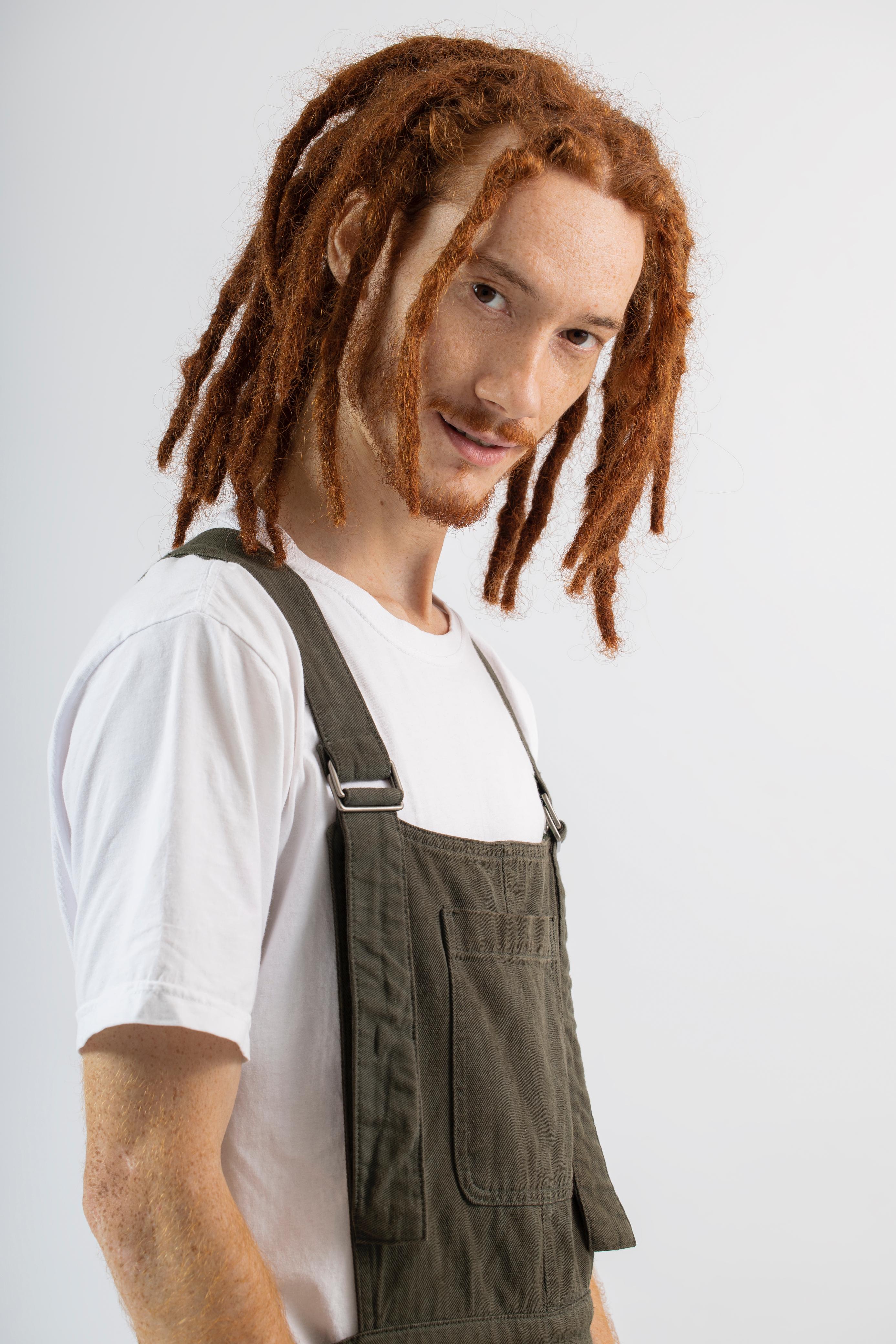 Retrato de Luan Paris, cabelos ruivos com dreads, camiseta branca e jardineira verde-musgo.